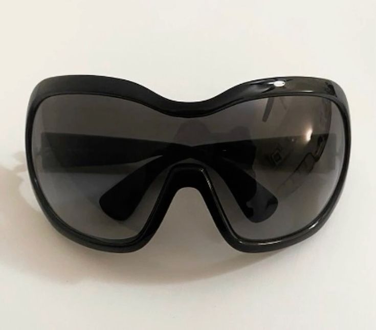 a closeup shot of a pair of black, oversized wraparound sunglasses. the sunglasses have a shieldlike shape and dark lenses.  they appear to be made of a glossy plastic material. the image is taken on a lightcolored surface.