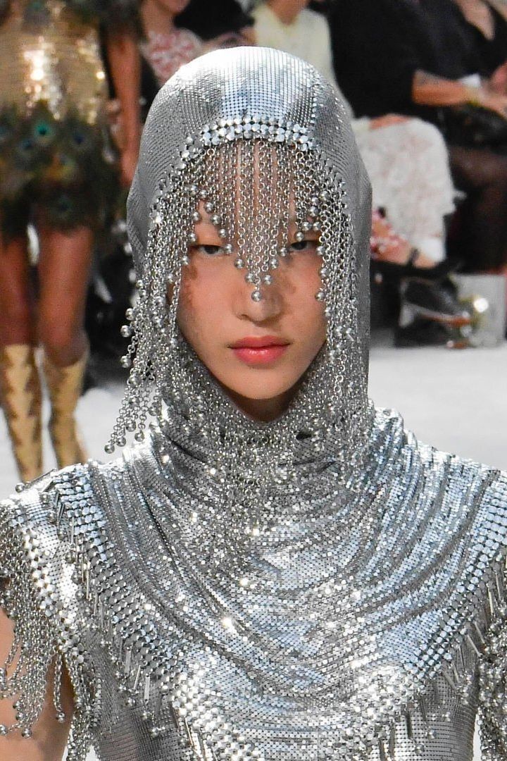 a woman in a silver chainmail dress and hood walks the runway at a fashion show. her hood is embellished with fringe and she is wearing a silver metal chain necklace. her makeup is simple with a bold red lip.