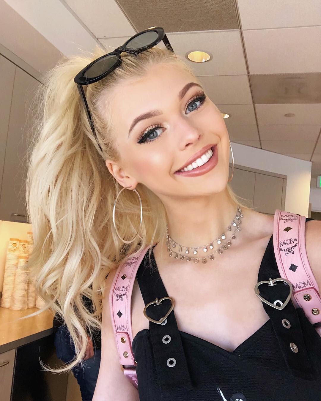 a woman with blonde hair pulled back in a ponytail, wearing sunglasses on her head, hoop earrings, a delicate necklace, and a black top with pink mcm backpack straps, is smiling and taking a selfie.