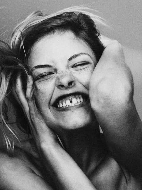 a black and white portrait of a young woman with her hand near her face, her mouth open in a wide smile,  showing her teeth. she has freckles and a missing tooth. her expression shows joy and happiness.