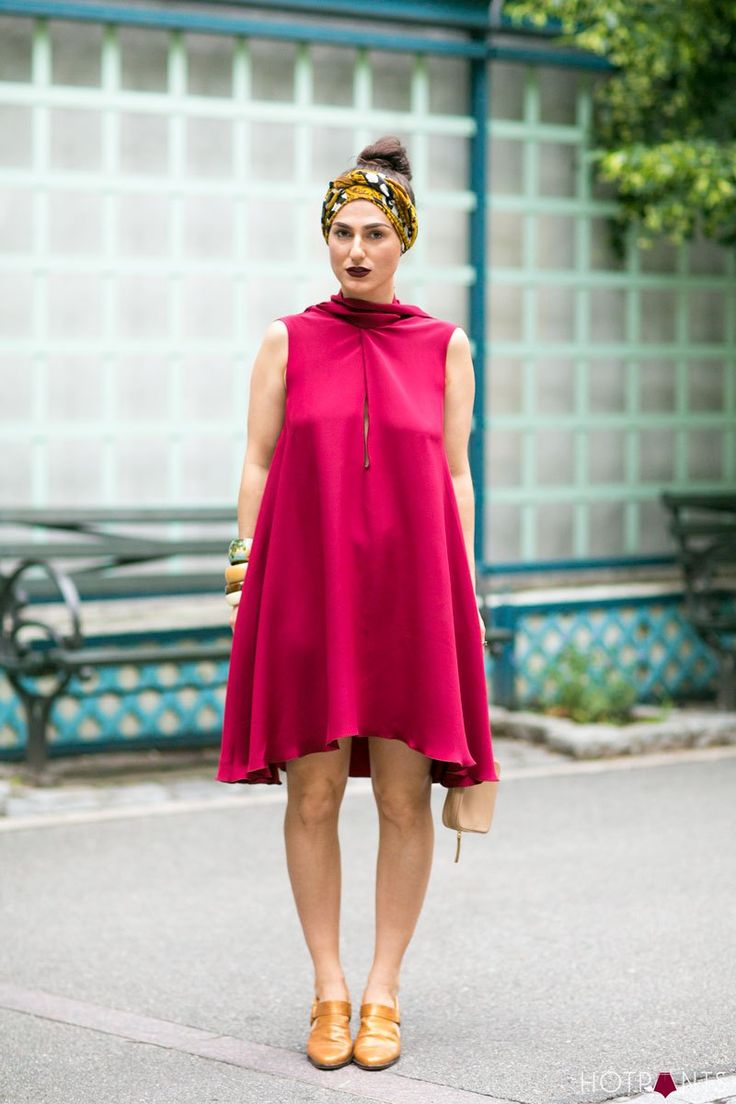 a woman is standing on a street wearing a red sleeveless tunic dress with a high neckline, a patterned head wrap, and brown leather shoes. she is also carrying a small beige handbag. the woman has her hair styled in a bun and is wearing a bracelet. she is standing in front of a background with a light blue wall and a green bench. the photo appears to be a street style fashion shot.