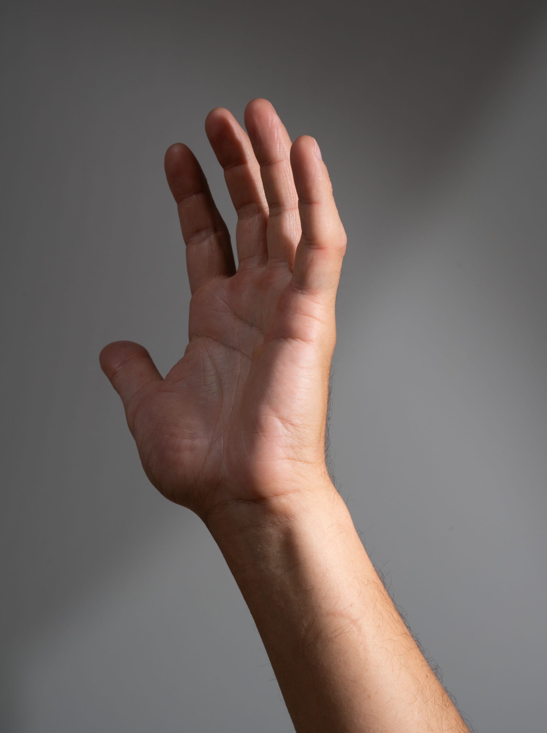 the image shows a man's hand reaching up against a gray background. the hand is welllit, with highlights and shadows that emphasize its form and texture. the fingers are slightly spread apart, and the palm is facing the viewer. the image has a sense of dynamism and movement, as if the hand is reaching for something just out of reach.