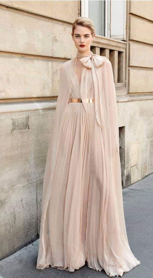 a full shot of a woman wearing a long, blush pink pleated gown with a capelike overlay. she is wearing a rose gold metallic belt and has her hair styled in a short, bob haircut. she is posing outdoors against the backdrop of a beige building with architectural details. the overall style is elegant and sophisticated.