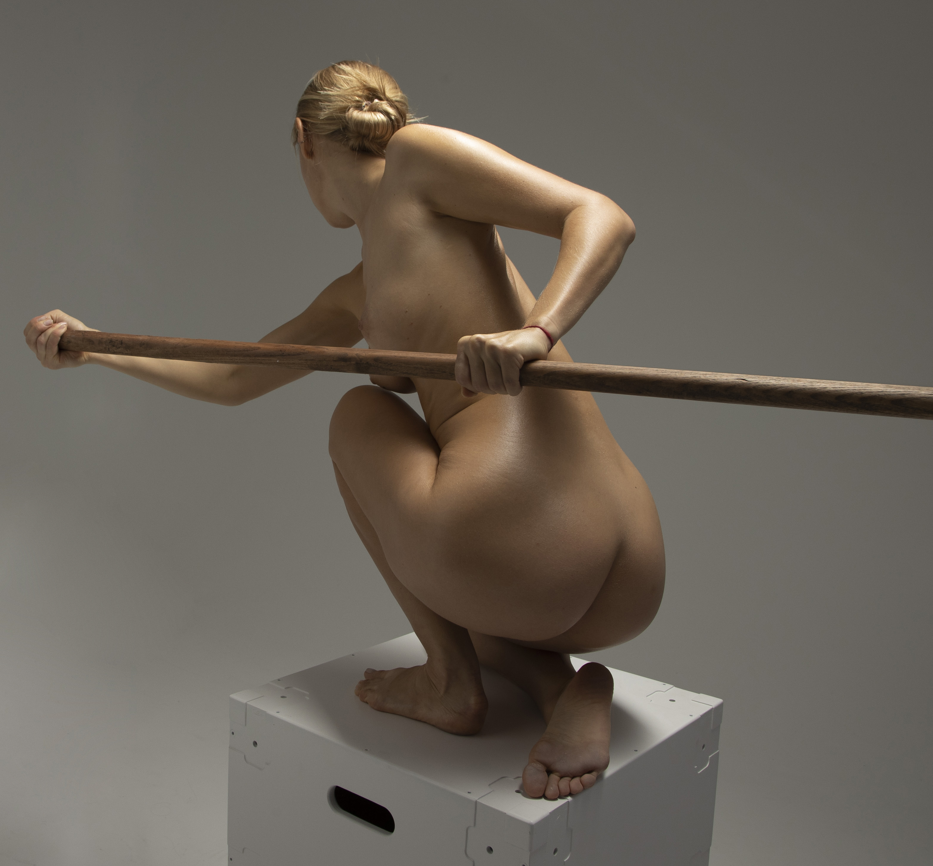 a nude woman with blonde hair in a bun is crouching on a white box in a studio setting. she is holding a wooden stick across her body. her back is facing the camera, and she is positioned against a gray background.