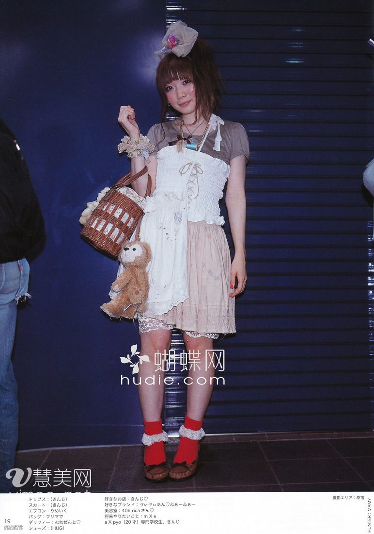 a young woman with brown hair in a small bun is standing indoors in front of a blue shutter door. she is modeling a white and beige dress with a lace trimmed apron, red socks, brown shoes, and a brown basket purse. she is also holding a teddy bear. there is japanese text on both sides of the image.