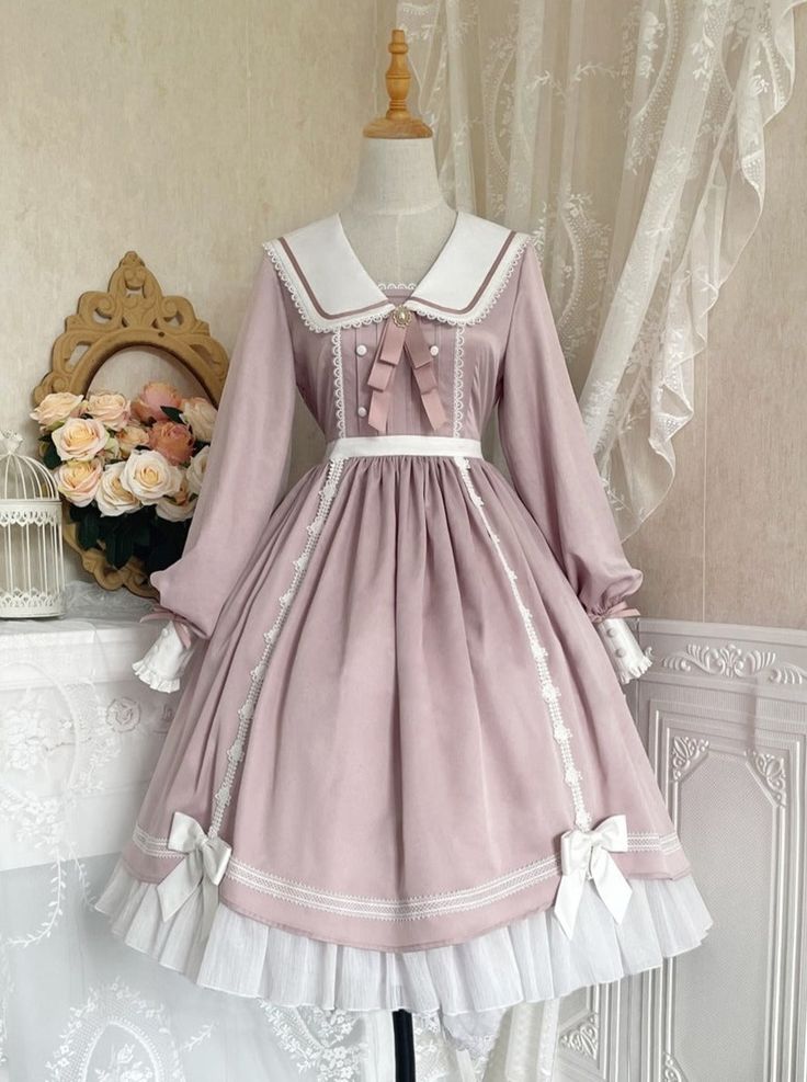 a pink lolita fashion dress with white trim is shown on a mannequin. the dress has long sleeves, a peter pan collar, and a bow at the neck. the skirt is full and has lace trim. the background is a white wall with a floral arrangement on the left side.