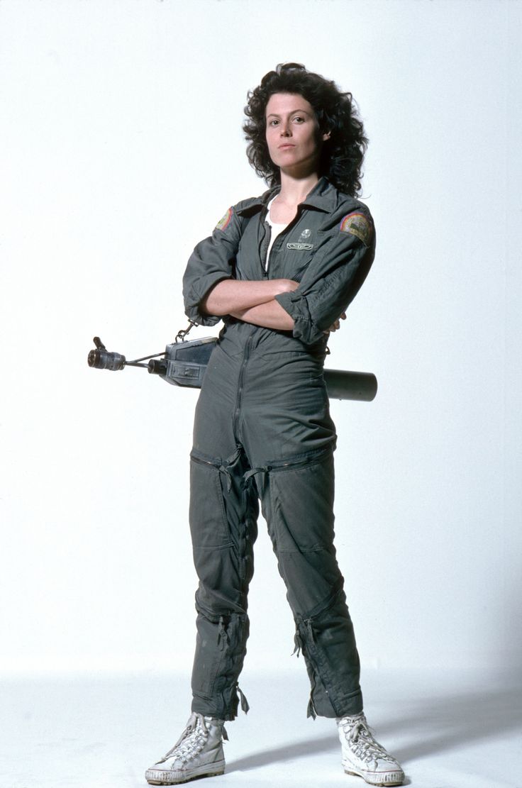sigourney weaver as ellen ripley, posing in a scifi jumpsuit, holding a gun in her right hand and looking into the camera. the background is white.  she is wearing white shoes.