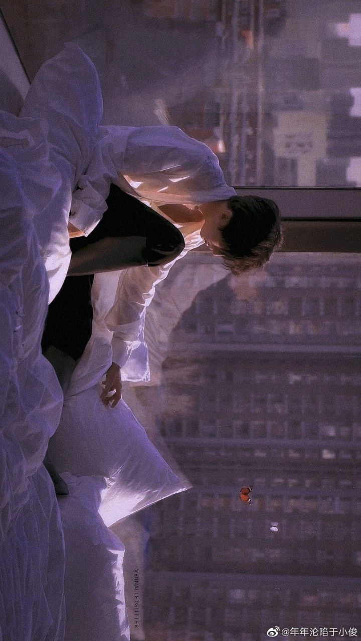 the image shows a young man sitting on a bed, his body leaning forward as he gazes out of a large window.  the perspective is from above, emphasizing the man's position within the vast cityscape visible beyond the window. the warm glow of sunset bathes the scene in a melancholic light. the overall impression is one of solitude and contemplation.
