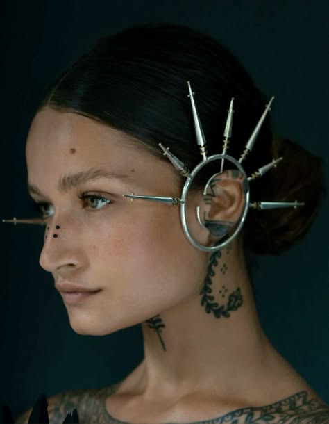 a close up portrait of a woman with dark hair wearing intricate silver jewelry, with a silver piece of jewelry attached to her ear and another silver piece connected to her nose. she is looking to the side and has tattoos on her neck and shoulder.  the background is dark.