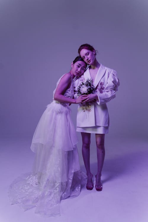 two women, possibly a lesbian couple, are captured in a studio shot against a purple backdrop. one woman is in a flowing white wedding dress with a long train, while the other is wearing a stylish white suit and short skirt. they embrace each other affectionately, holding a bouquet of flowers. the image exudes elegance and romanticism, suggesting a wedding or celebratory event.  the overall mood is soft and intimate, highlighting the couple's love and connection.