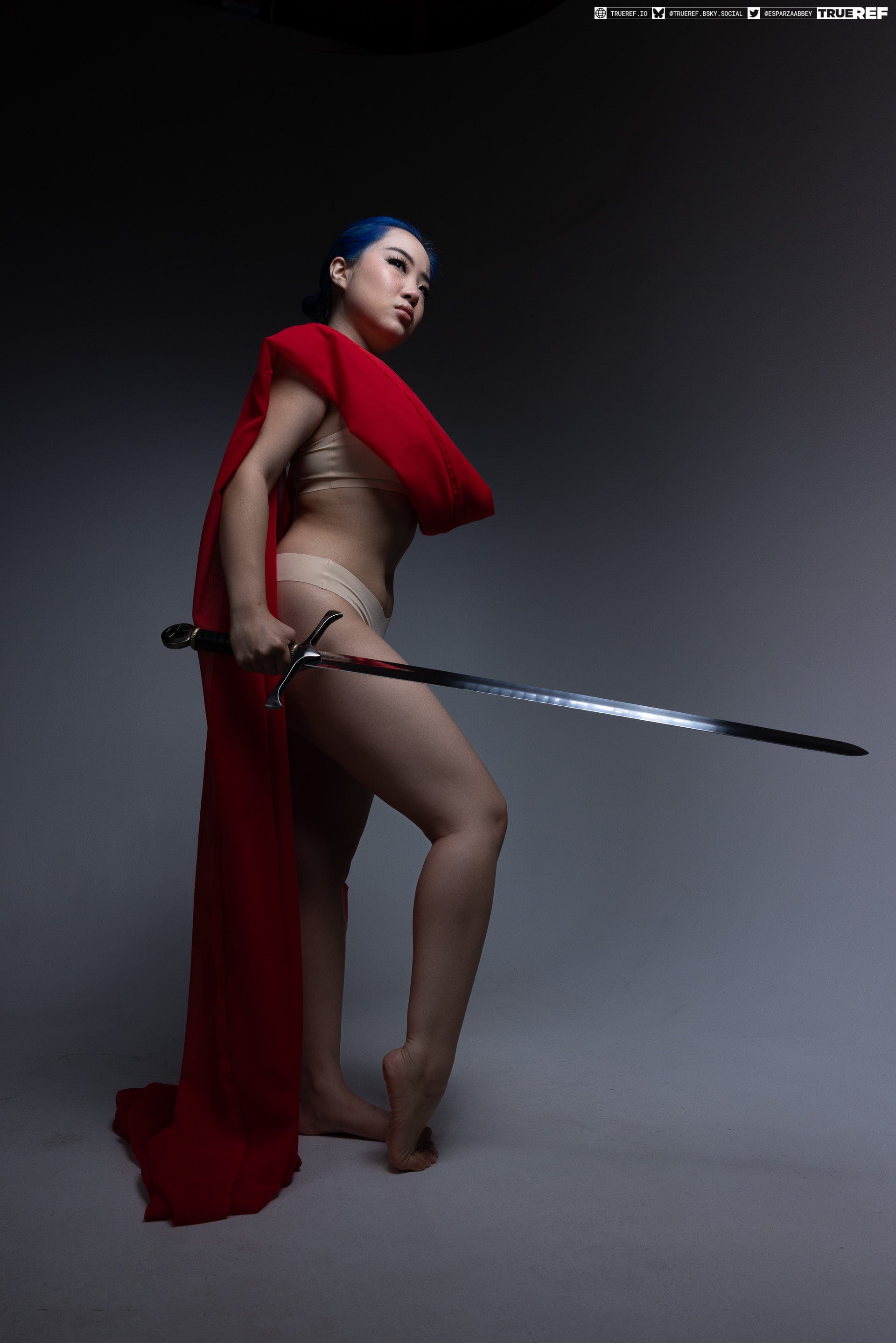 a woman with blue hair stands in a studio setting, holding a sword. she is wearing minimal clothing and a red cape. she is looking up and away from the camera. she is standing on bare feet.