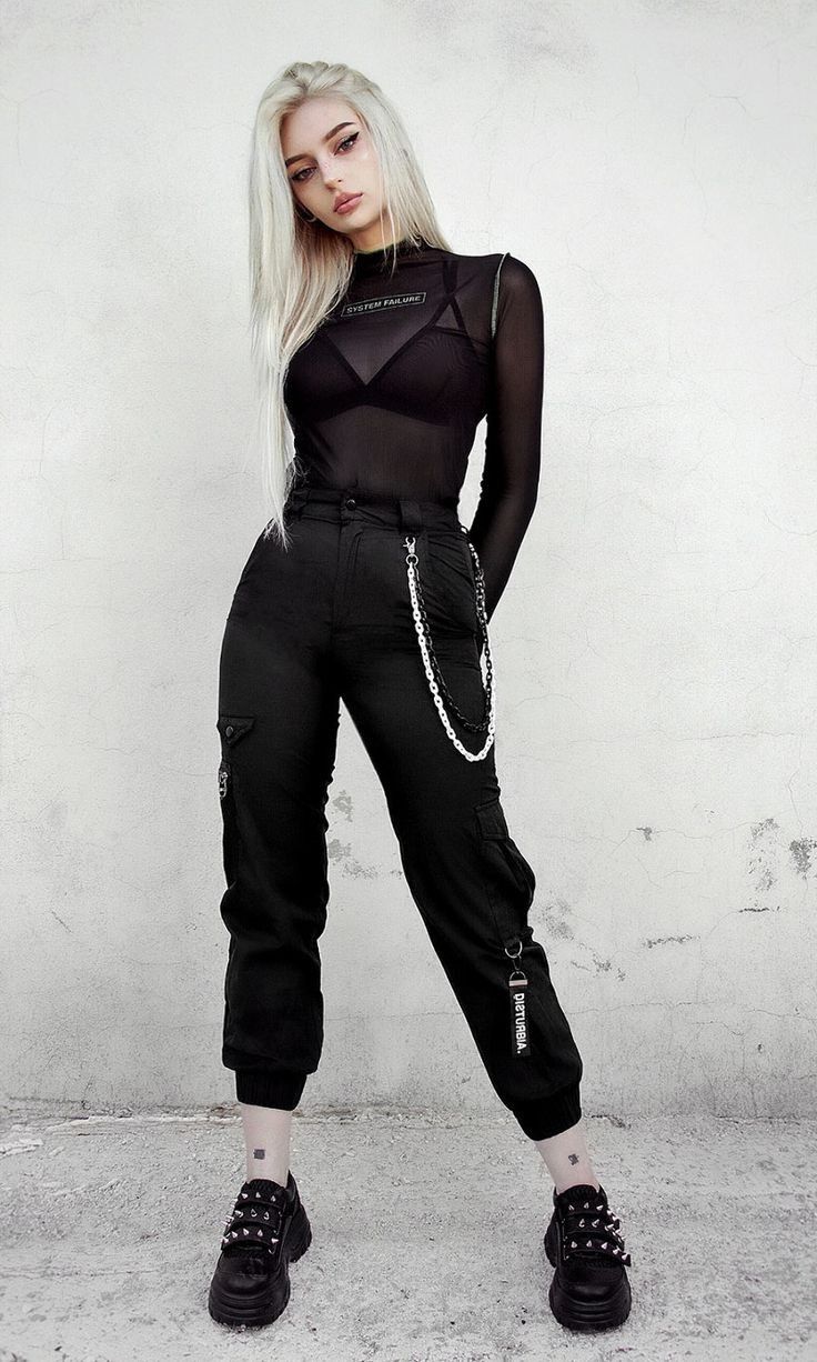 a fullbody shot of a blonde woman wearing a black sheer longsleeve top, black cargo pants, and black studded boots. she is posing against a grey wall. her style is dark, edgy, and alternative.