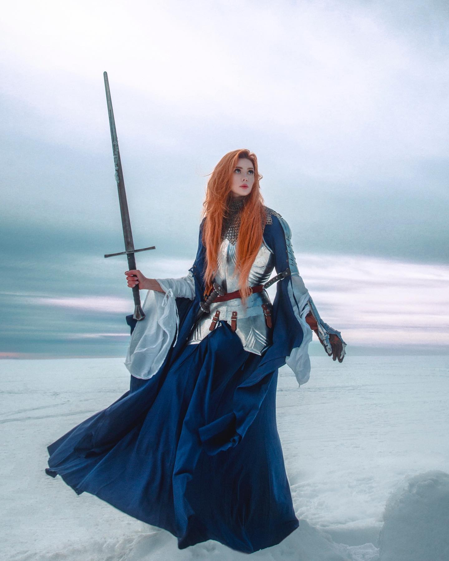 a full shot of a woman cosplaying as a medieval knight. she is wearing a blue dress over shining silver armor and carrying a long sword. her long red hair flows down her back, and she is standing in a snowy landscape, looking off into the distance. the scene is evocative of a fantasy setting. the overall impression is one of strength, beauty, and a sense of quiet power.