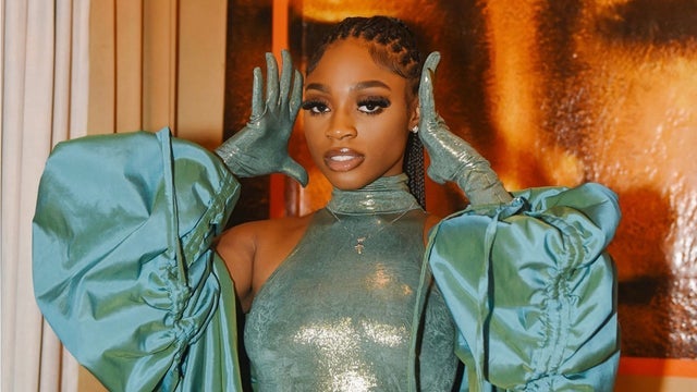 a young black woman with long braided hair in a teal green and gold shiny  outfit and matching teal gloves poses for a photo with her arms raised above her head.