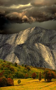 a dramatic landscape with a large mountain in the background and a valley in the foreground. the mountain is covered in rocks and has a dark, cloudy sky above it. there is a lot of green grass in the valley and some trees. the landscape is very scenic and natural.