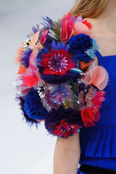 loseup view of a woman's shoulder adorned with a stunning floral embellishment. the embellishment is a vibrant mix of colors, including blues, reds, pinks, and purples, and features numerous textured fabric flowers and elements. it's attached to a bright blue dress, suggesting a highfashion or couture piece. the image focuses on the intricate detail and artistry of the floral design, showcasing the texture and variety of fabrics used in its creation. the woman's arm and shoulder are visible, partially obscured by the elaborate embellishment. the background is a plain, lightcolored surface.