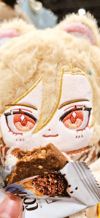 a kawaii plush toy with brown hair and big eyes is holding a chocolate cookie.  the plush is holding the cookie in the air in its tiny hands.  the plush is sitting on a white background.