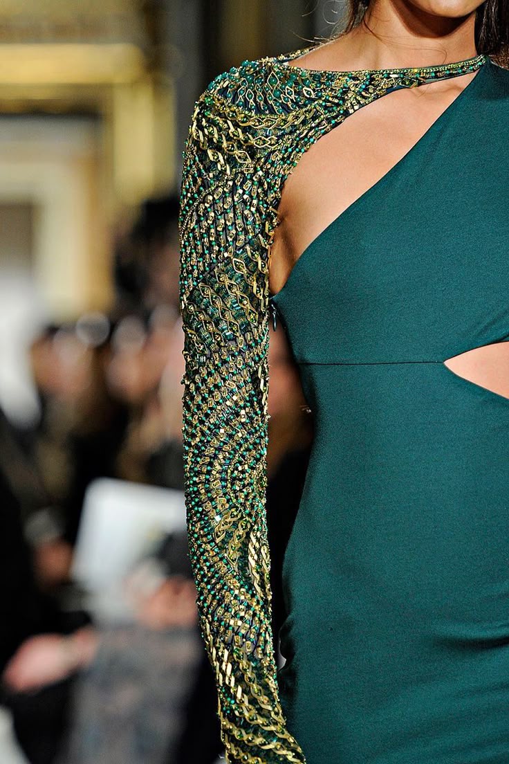 closeup of an emerald green dress featuring a long, heavily embellished sleeve. the dress has a cutout design and a oneshoulder neckline. the embellishments include beads and goldcolored details.