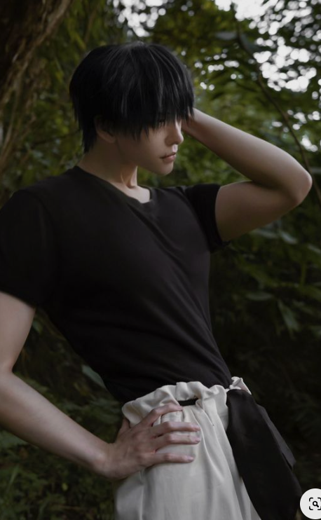 a man cosplaying an anime character with black hair is posing in a forest. he is wearing a black tshirt and white pants with a black sash. the lighting is natural and diffused, creating a soft and ethereal look. the perspective is eye level, giving the viewer a sense of intimacy with the subject.