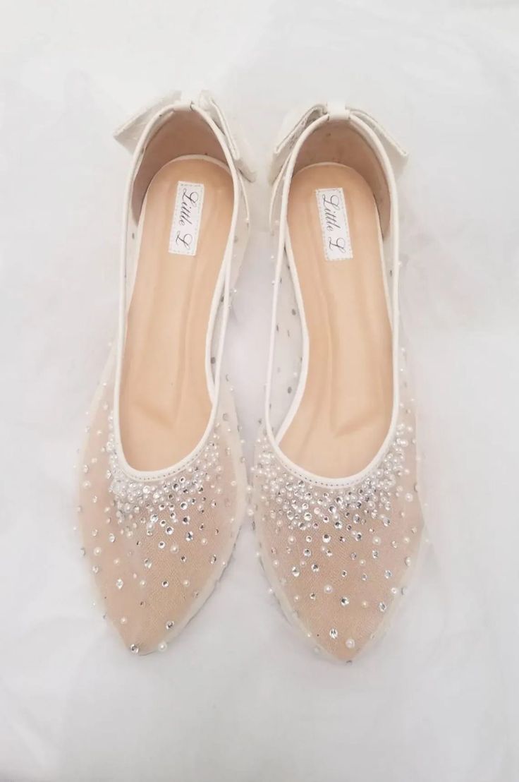 a pair of white bridal shoes with lace and rhinestones. the shoes are flat and have a pointy toe. the shoes are made of white satin and have a white lace overlay. the lace is embellished with rhinestones. the shoes have a bow on the top.  the shoes are perfect for a wedding or any special occasion.