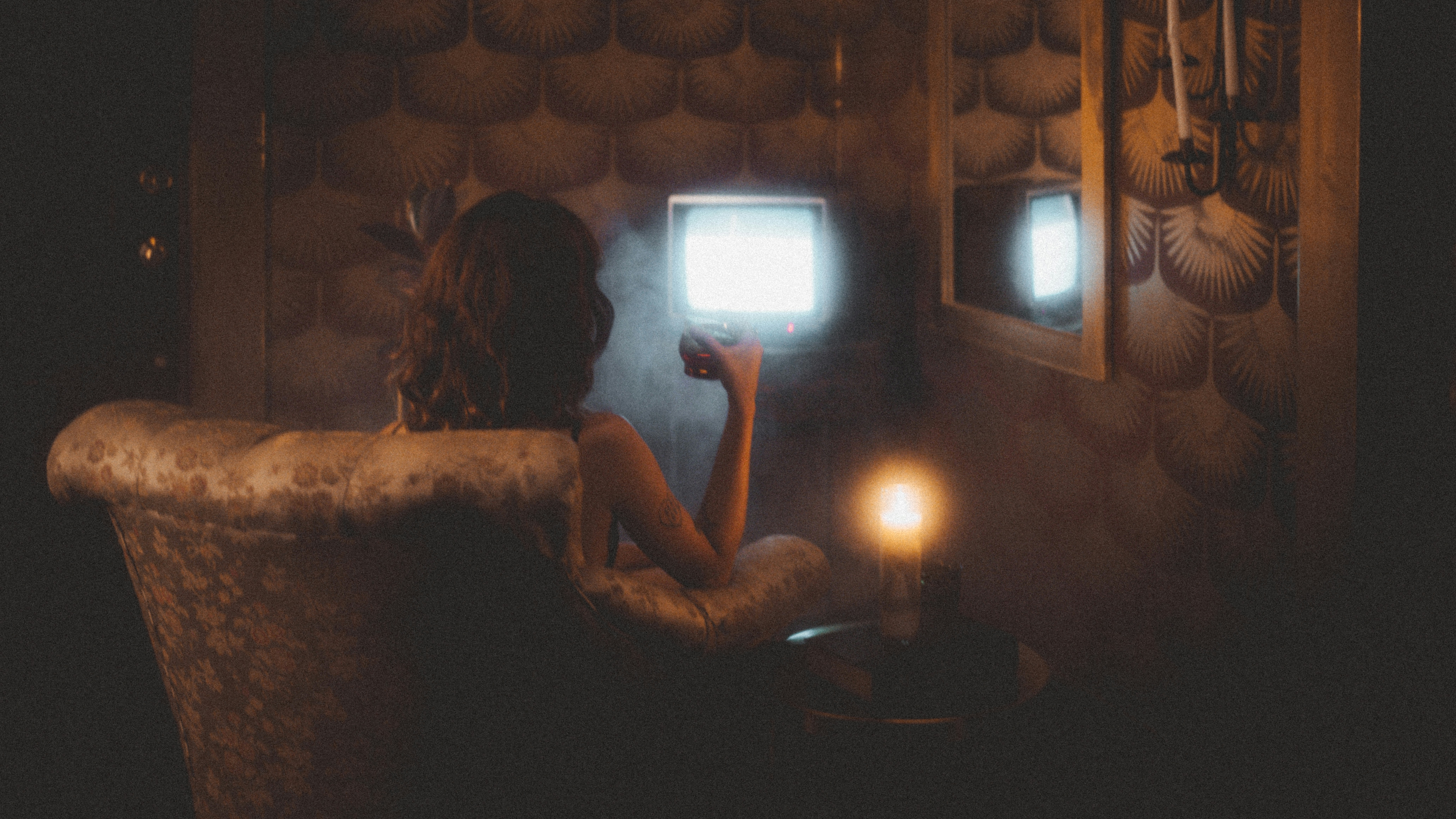 a woman is seated in a plush armchair, her back to the viewer, facing two vintage televisions that illuminate the dimly lit room. the warm glow of a flickering candle on a nearby table adds to the cozy and somewhat mysterious atmosphere. the room is decorated with patterned wallpaper, hinting at a retro aesthetic.