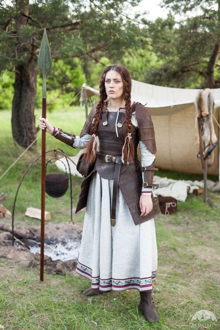 a woman dressed in medieval armor stands outdoors, holding a spear. she has braided hair and a serious expression. behind her is a campsite with a tent, a cooking pot over a fire, and trees in the background. she is wearing a long dress, leather armor, and boots. the scene appears to be a historical reenactment or a staged photograph.