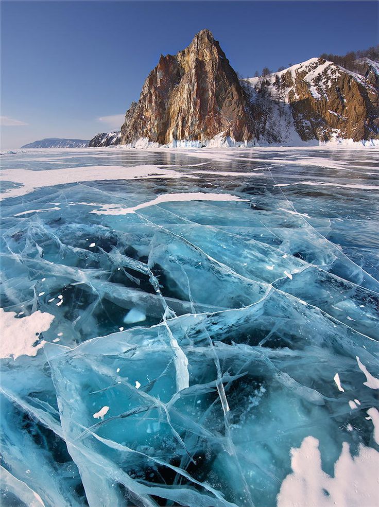 a stunning landscape featuring the frozen surface of lake baikal in siberia, russia, during winter. the foreground showcases cracked and clear blue ice, with patches of snow scattered across the surface. in the background, a rocky mountain with snowcovered peaks rises under a clear blue sky. the scene captures the natural beauty and cold environment of the region.