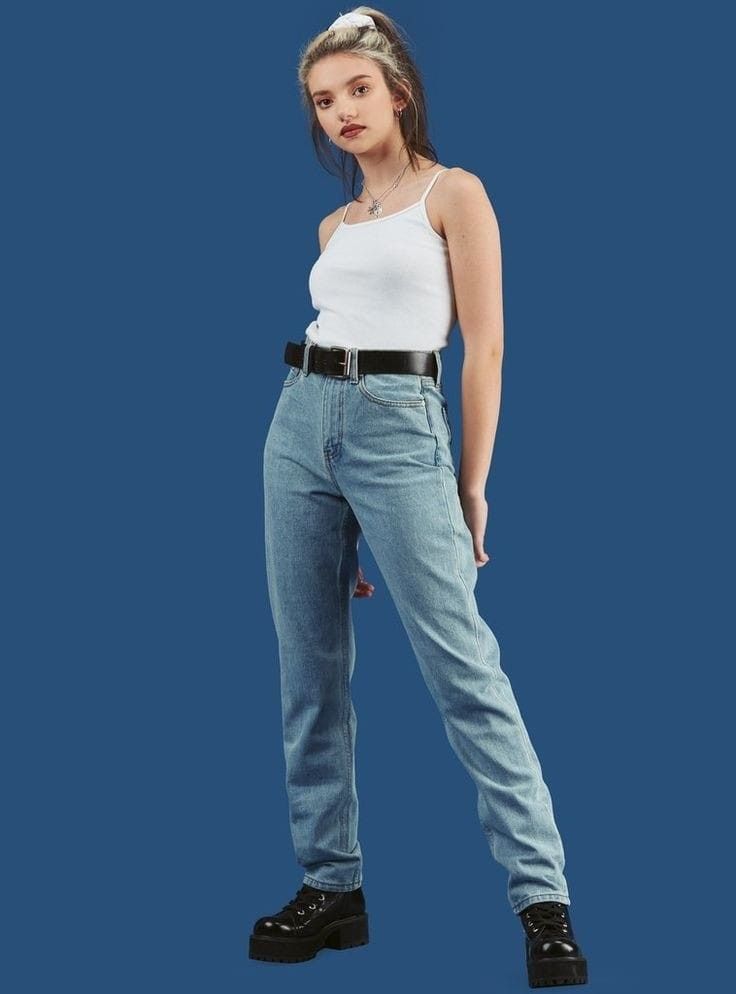 a fullbody shot of a young woman model posing against a solid blue background. she's wearing light wash straightleg jeans with a high waist, a simple white tank top, a black belt, and black chunky platform boots. her hair is styled up in a halfponytail. the overall style is casual and reminiscent of 90s or grunge fashion.  the image is welllit and professionally styled.