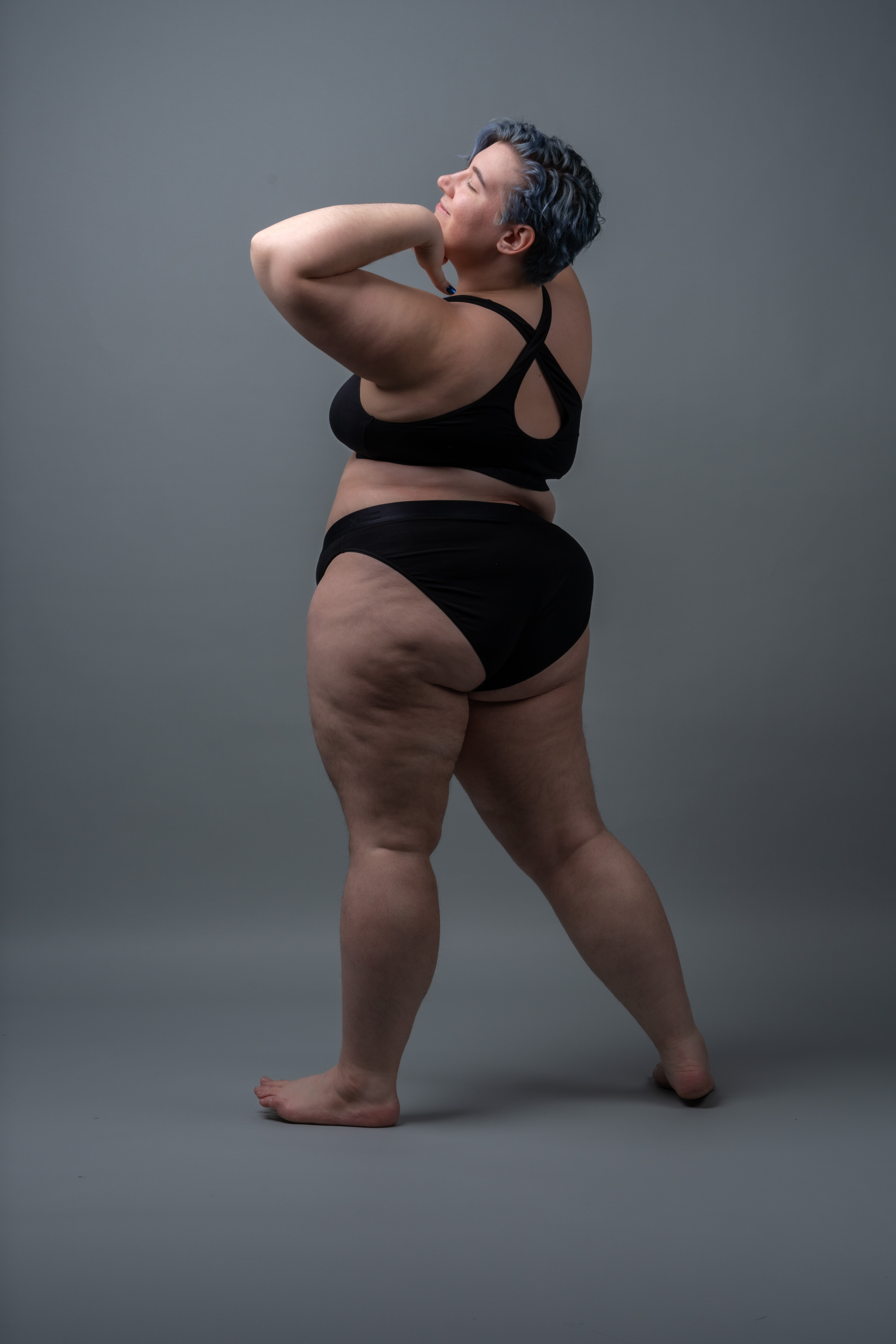 a fullbody studio shot of a plussize woman with blue hair, posing in black underwear against a grey background. she has her arms raised and is looking upwards, showing her natural skin and cellulite. she is barefoot.