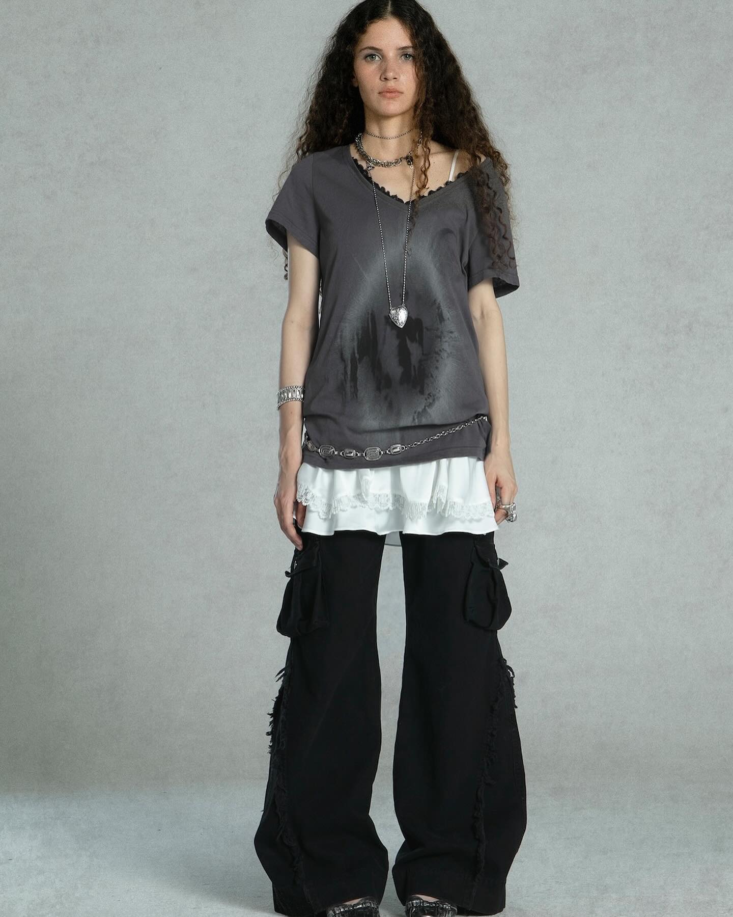 a fullbody shot of a woman model showcasing a stylish outfit. she is wearing a grey, distressed vneck tshirt layered over a white lace trim top, paired with black wideleg cargo pants with frayed detailing at the hems.  the model wears multiple necklaces, bracelets and rings, adding to the grunge aesthetic. the backdrop is a neutral grey, keeping the focus on the clothing.