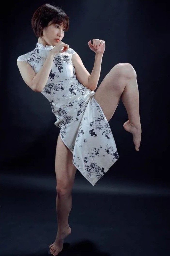 a woman in a white and black floral cheongsam stands on a dark background in a martial arts pose.  she is barefoot and looking off to the side, her expression focused and alert. her right leg is raised high in the air, showcasing her flexibility and strength. the lighting is dramatic, highlighting the contours of her body and the flowing movement of the dress. the overall image conveys a sense of power, grace, and cultural pride.