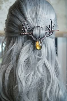 a woman with long, silver hair is wearing a unique antler hairpin. the hairpin is a intricate design with a  large, ambercolored stone in the center. the hairpin is positioned in the woman's hair, holding her hair back from her face. the woman has a beautiful, natural look and her hair is styled in a simple, elegant way. the hairpin adds a touch of bohemian style to her look.