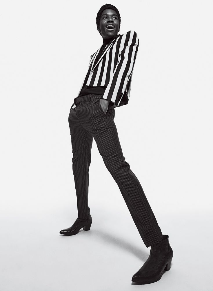 a young man wearing a striped jacket and pinstripe pants is standing in a studio. he is posing and smiling at the camera. he is wearing a black turtleneck and black boots. he is looking off to the side. the background is white.