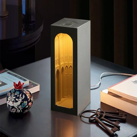 a modern lamp shaped like a church is sitting on a table next to a few books and keys. the lamp is lit, and the light is casting a warm glow on the surrounding objects.