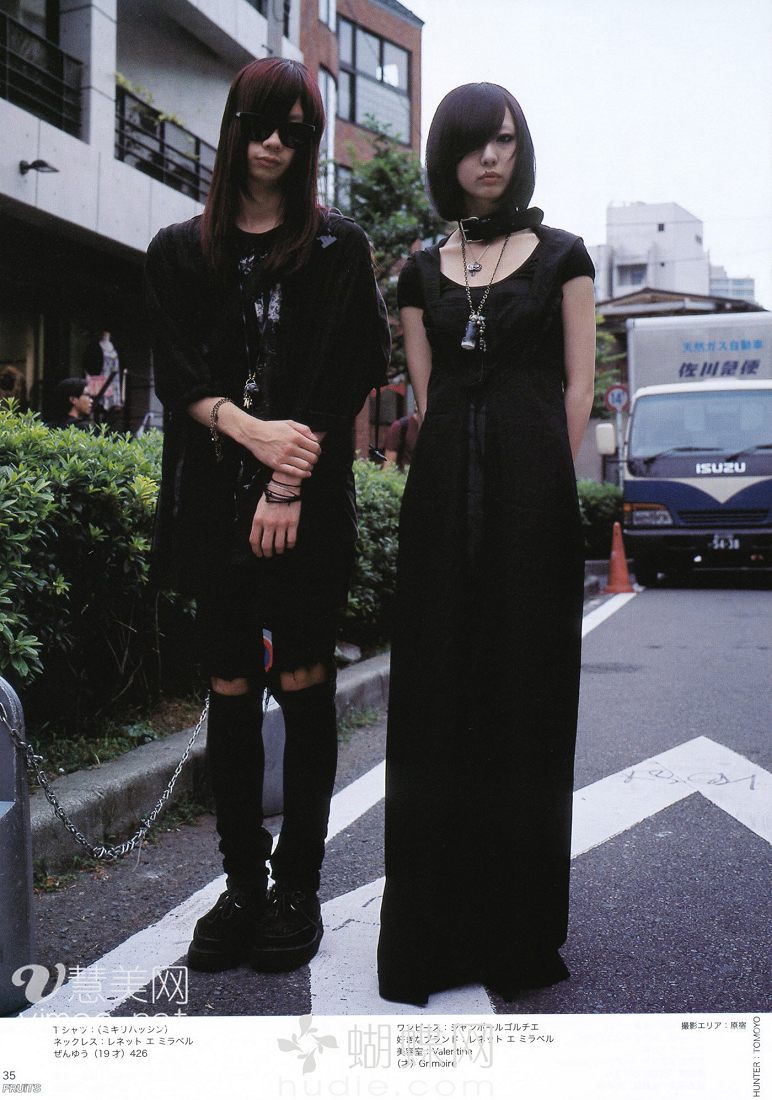 two young adults are posing on a street, showcasing their unique fashion styles. both are dressed predominantly in black, with the woman wearing a long, flowing dress and the man sporting ripped jeans and a tshirt. they accessorize with silver jewelry and dark sunglasses, giving off a visual kei or gothic vibe. the man stands with his arms crossed, while the woman keeps her hands by her side. their expressions are serious and composed. the setting appears to be a typical japanese street, with buildings and a parked truck in the background. the photo is taken in natural daylight, casting a bright yet slightly muted light on the scene.