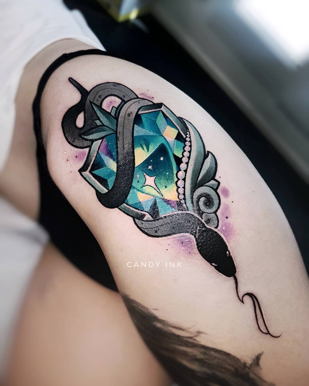 a tattoo of a snake wrapping around a jewel on someone's upper thigh. the tattoo is done in a neotraditional style with black linework and watercolorlike shading.