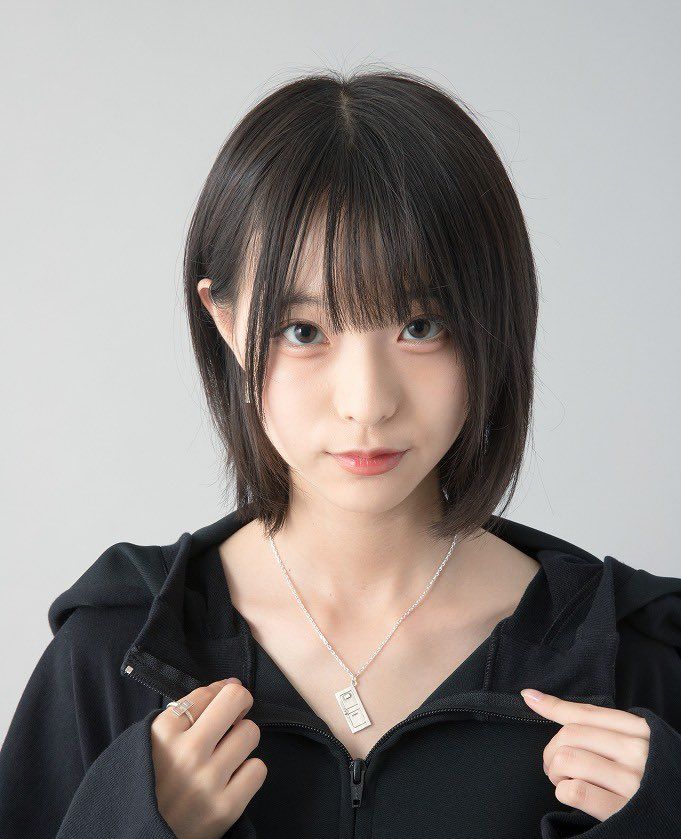 a young asian woman with black hair, wearing a black hoodie and a silver necklace, is looking at the camera with a slight smile. she is standing against a white background in a studio setting.