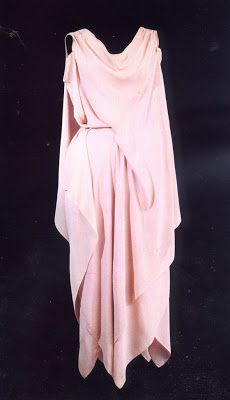 the image shows a pale pink, sleeveless dress. the dress is draped and folded in a way that creates a flowing and elegant look. it has a cowl neckline and appears to be made from a lightweight fabric. the style is reminiscent of 1930s fashion or a grecianinspired design. the dress is displayed against a dark background, emphasizing its light color and flowing lines.