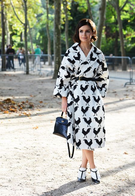 a stylish woman strolls down a parisian street, showcasing her chic autumnal ensemble. she's wearing a kneelength coat adorned with a striking rooster print in black and white. the coat is belted at the waist, accentuating her figure.  she carries a structured black handbag and sports elegant black and white ankle strap shoes. the background features a treelined avenue, suggesting a relaxed yet sophisticated atmosphere. the overall impression is one of effortless parisian style.
