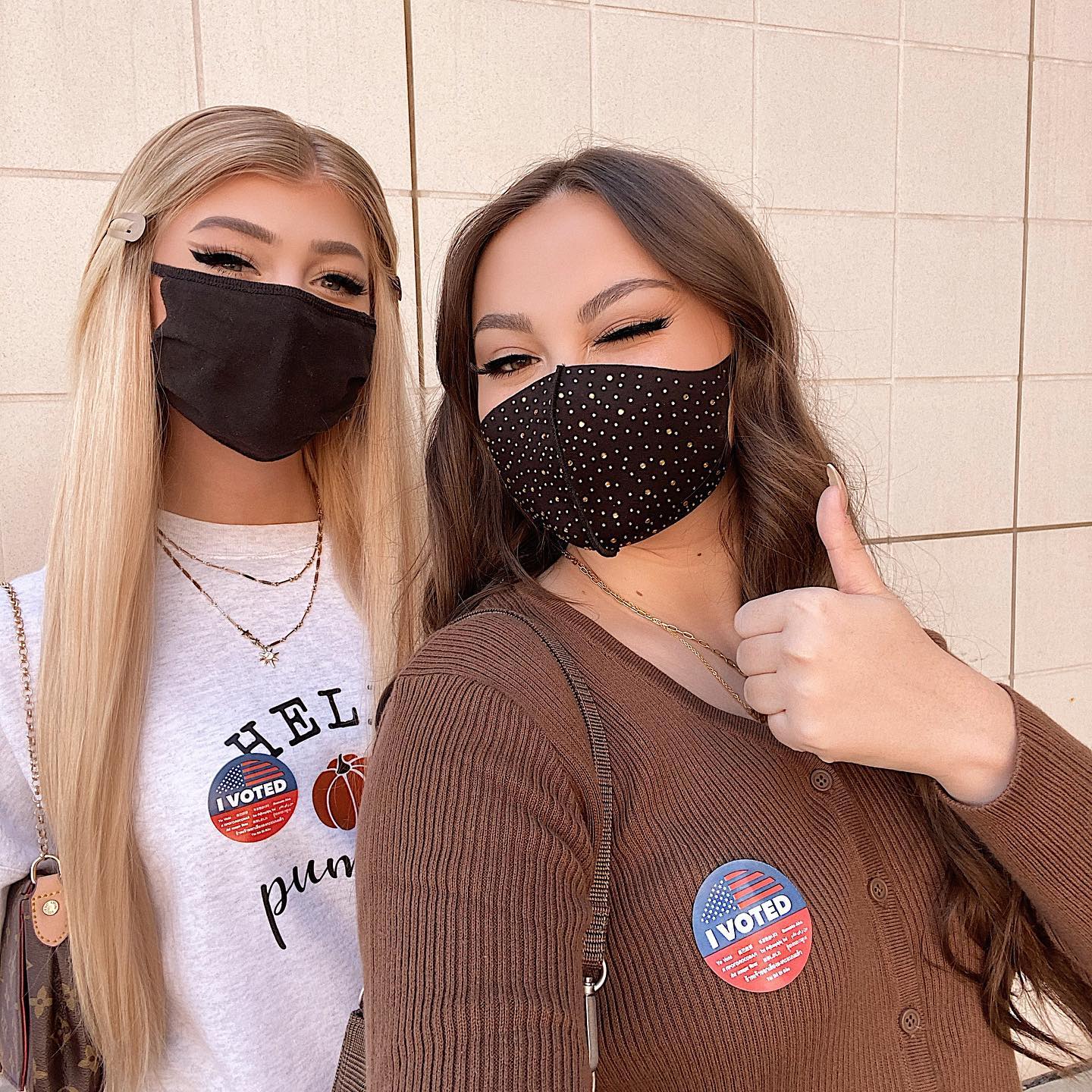 two women wearing face masks stand sidebyside. the woman on the left has blonde hair, a black face mask, and a white sweatshirt with an "i voted" sticker. the woman on the right has brown hair, a black face mask with white dots, and a brown cardigan. she is giving a thumbsup and also has an "i voted" sticker on her cardigan. both women have necklaces and purses. they are standing in front of a lightcolored wall.