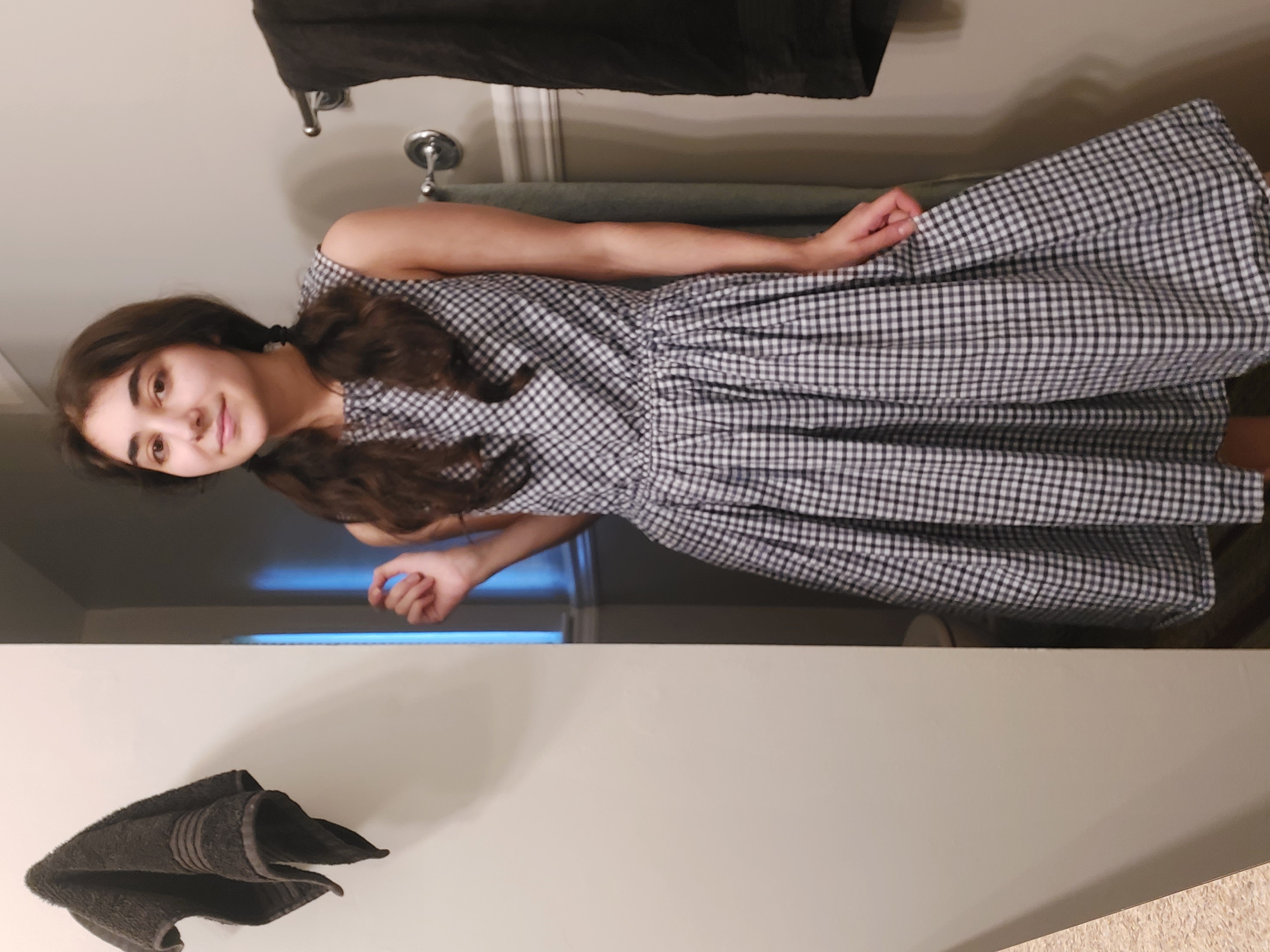 a young woman with pigtails stands in a bathroom, wearing a blue and white gingham midi dress. she is positioned slightly angled, her body facing towards the right of the frame.  the dress is sleeveless and has a gathered waistline. the bathroom setting is minimalist with a white wall and a darkcolored towel hanging on a rack. a darkcolored towel is also on the wall in the background.  the woman appears to be posing for a photo.