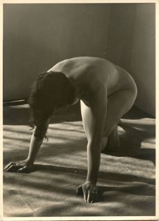 a black and white photograph of a nude woman kneeling on the floor.  the woman's body is arched, her head and upper body bent forward, and her arms extended towards the ground. strong shadows and light contrast highlight the curves of her body. the photo appears to be vintage or retro in style. the setting is a simple, indoor studio space.