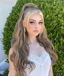 a young woman with blonde and brown wavy hair styled in a high ponytail stands outdoors in front of a green bush. she is wearing a white top with lace detailing and gold hoop earrings. she is wearing makeup including eyeliner.