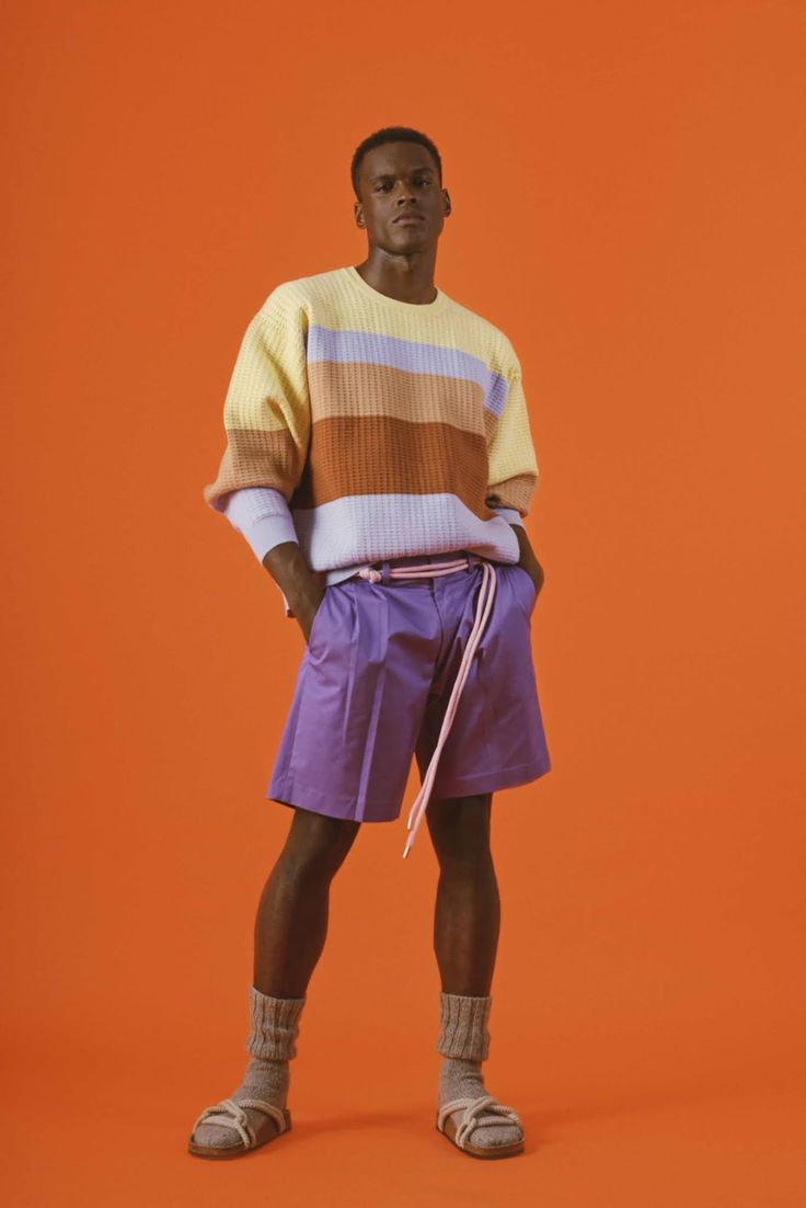 a full shot of a darkskinned male model standing against a bright orange background. he's wearing a pastelcolored horizontally striped sweater, purple shorts, light gray socks, and sandals.  the model is casually posing with his hands in his pockets.  the overall style is casual and summery.
