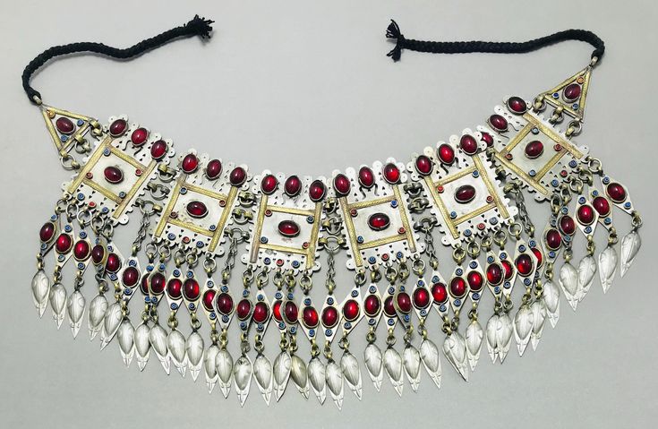 a closeup studio shot shows a vintage ethnic silver and gold choker necklace with red gems and black cord. the necklace features square and teardrop pendants.