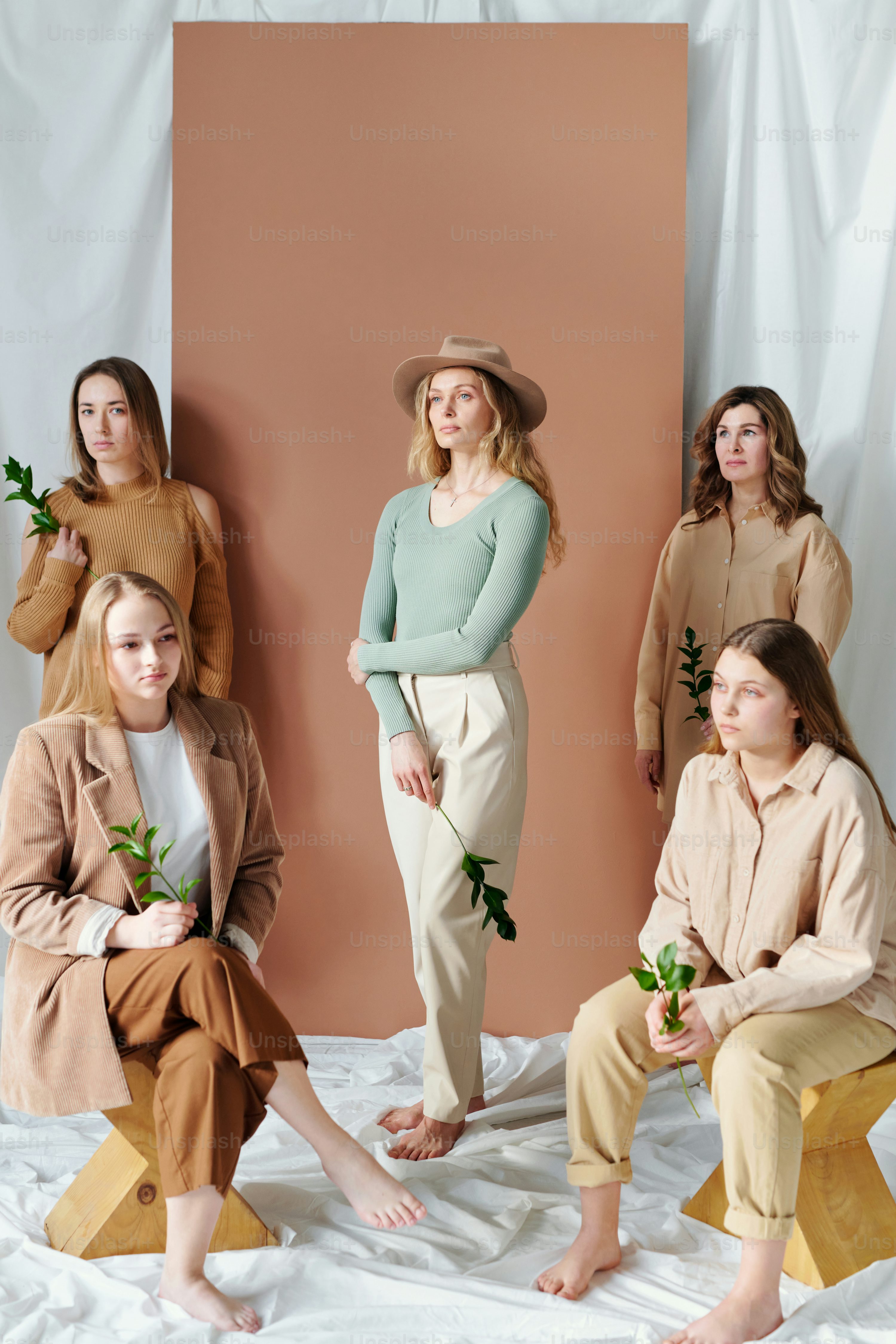 ive women of different ages are posed in a studio setting against a light brown backdrop. they are dressed in various shades of beige and brown clothing, creating a cohesive and stylish aesthetic.  some are sitting on wooden stools, others standing.  they each hold small green sprigs, adding a natural element to the image. the overall mood is calm and relaxed.  the women appear to be related, possibly sisters or close friends, creating a sense of generational connection.