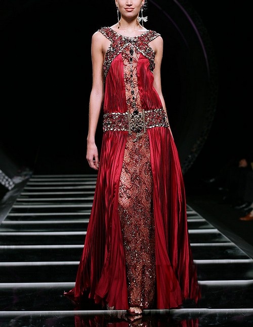 the image showcases a model on a runway wearing a stunning, floorlength red gown. the dress is heavily embellished with beading and sequins, particularly at the neckline and waistline, creating a luxurious and vintage look reminiscent of the 1920s flapper era. the dress features a pleated skirt and a fitted bodice. the model is elegantly posed, and the dark background highlights the dress and the model’s beauty. the overall aesthetic is one of high fashion and sophistication.
