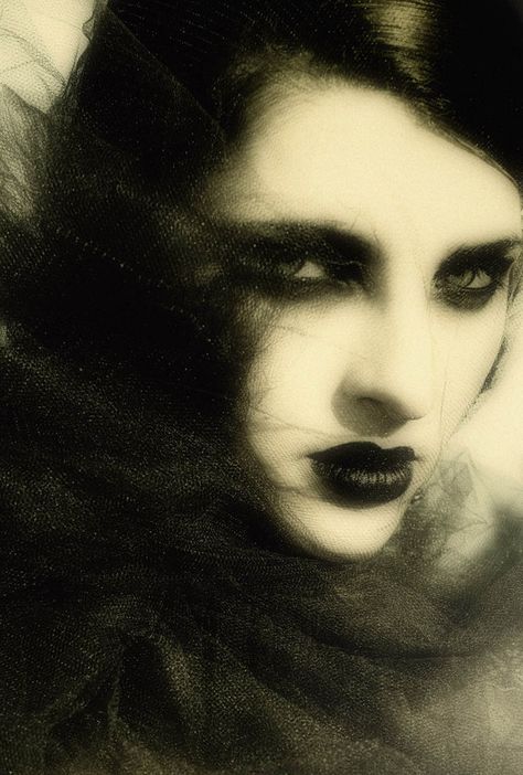 a closeup portrait of a woman with dark hair and makeup, her eyes looking intensely at the camera. her lips are painted a dark red. she is partially obscured by a black veil, which adds to the mysterious and seductive atmosphere of the image. the overall tone of the image is dark and gothic.