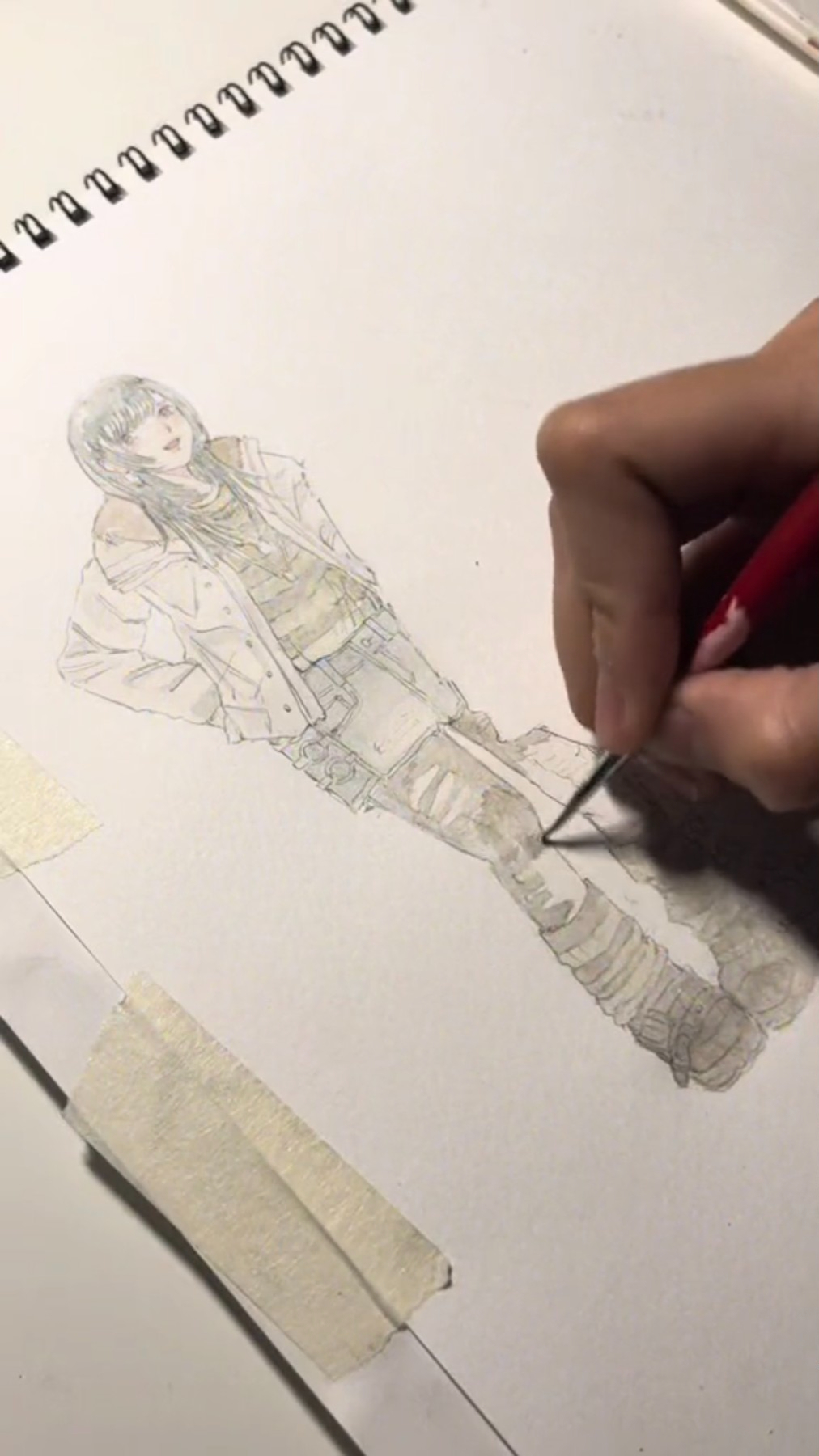 a person's hand is drawing a female figure on a white piece of paper. the drawing is in pencil and is of a girl wearing a white jacket with a black and white shirt underneath.  the figure is wearing ripped pants and what looks like boots on her feet.  the artist is adding details to the image by using a pencil. the background of the image is blurred.