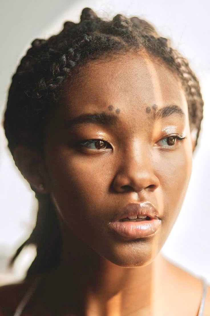 a closeup portrait of a young woman with dark brown skin and braided hair, looking to the right. she has brown eyes, glossy lips, and distinctive dark markings on her forehead between her eyebrows. sunlight casts a shadow on part of her face. the shot is framed from the head and shoulders.